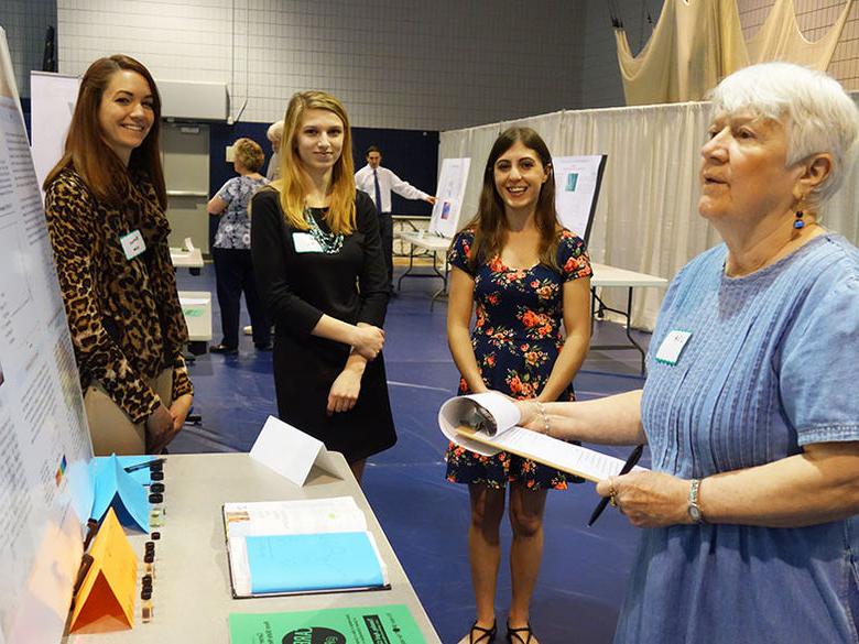 A judge reviews the poster presentation of three female students at the Research and Creative Exposition